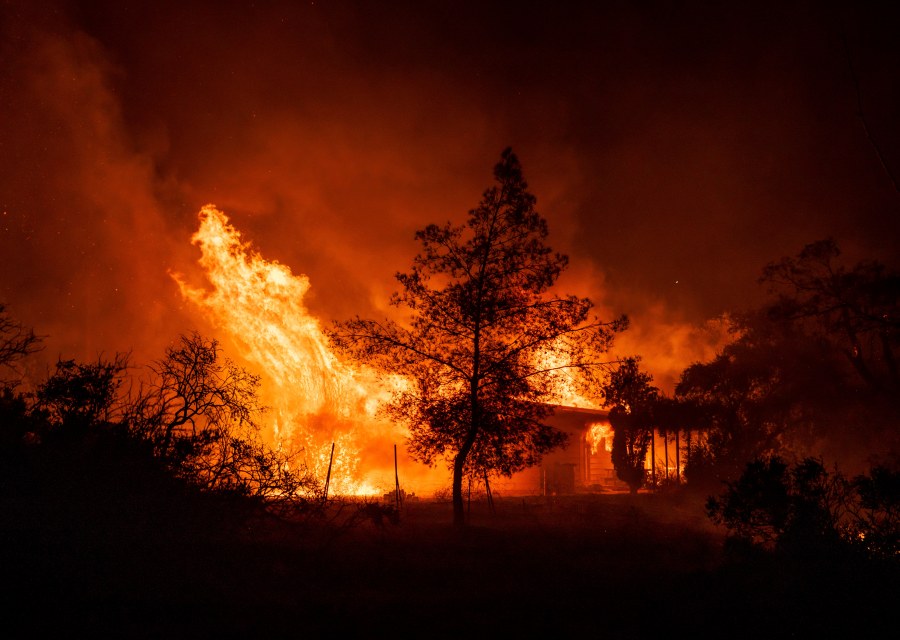 A structure is engulfed as bright red flames from a wildfire flare against a dark sky.