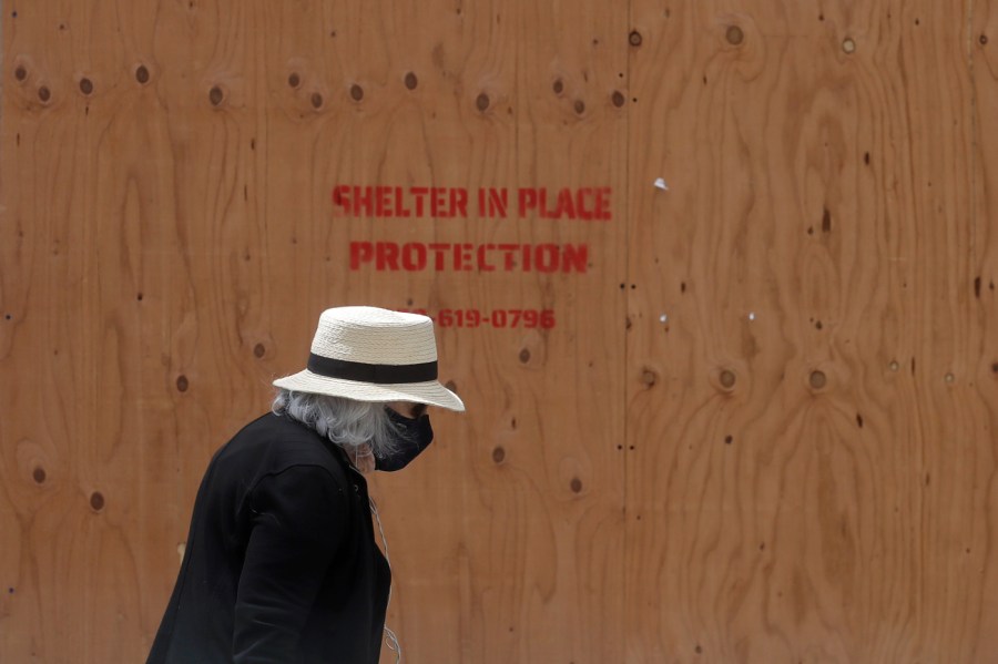 A woman wears a face mask while walking past boarded windows at a store in San Francisco, Saturday, April 18, 2020. (AP Photo/Jeff Chiu)