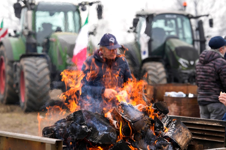 A farmer sits next to a fire during a gathering near the highway junction in Melegnano, near Milan, Italy, Wednesday, Feb. 7, 2024. Farmers have been protesting in various parts of Italy and Europe against EU agriculture policies. (AP Photo/Luca Bruno)