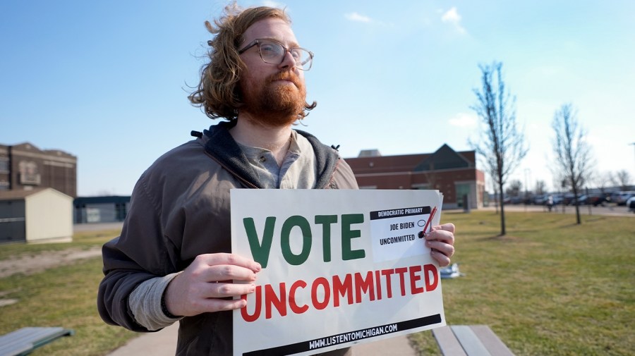 A demonstrator holds a "Vote Uncommitted" sign outside a voting location.