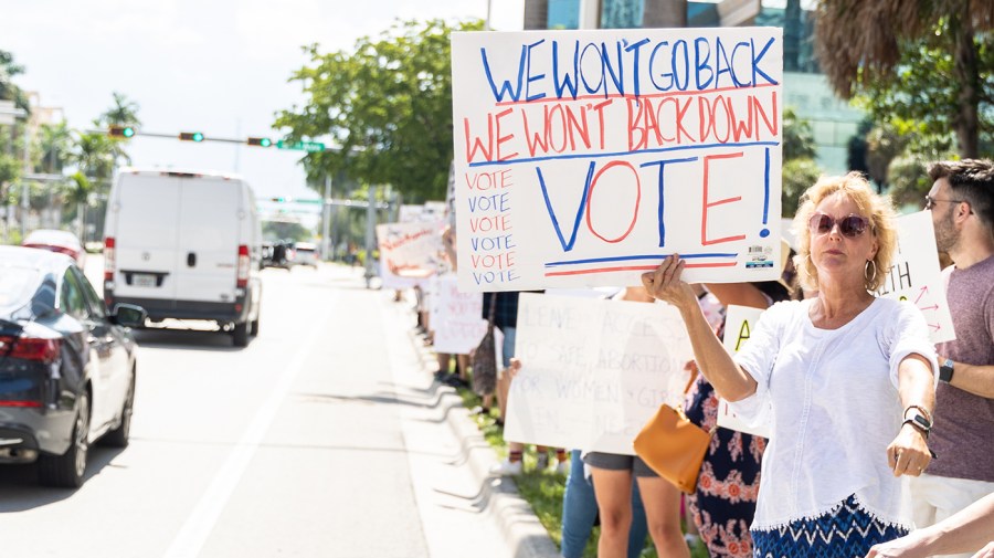 Protesters hold signs along a roadway. One sign reads, "We won't go back. We won't back down. Vote!"