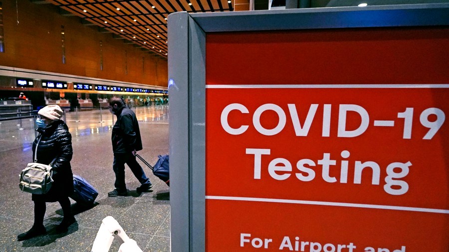 FILE - Travelers pass a sign near a COVID-19 testing site in Terminal E at Logan Airport, on Dec. 21, 2021, in Boston. The nation’s top public health agency is expanding a program that tests international travelers for COVID-19 and other infectious diseases. The Centers for Disease Control and Prevention currently operates a program at six U.S. airports that asks passengers from inbound international flights to agree to nose swabs and answer questions about their travel. (AP Photo/Charles Krupa, File)