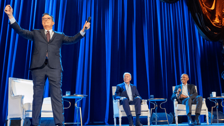 Stephen Colbert speaks with his hands up in the air, as President Biden and former President Barack Obama watch in the background.