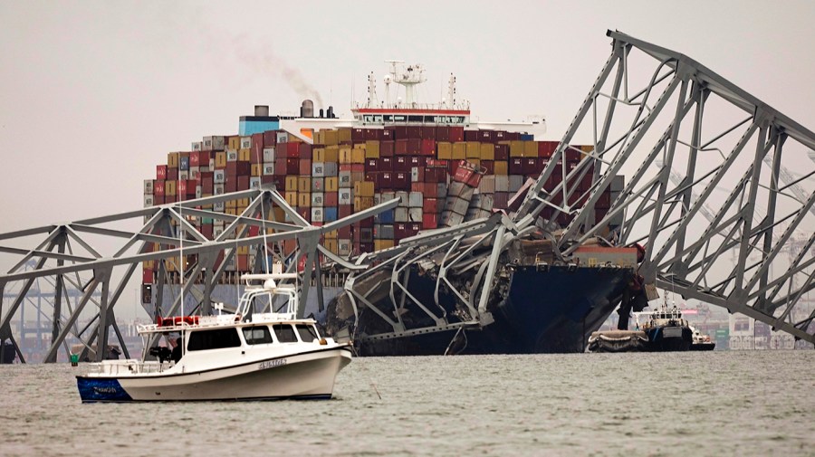 A small boat is anchored in front of a cargo ship stuck under the part of the structure of the Francis Scott Key Bridge.