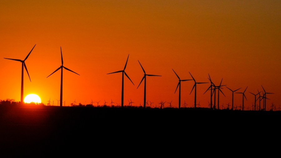 The setting sun lights up an orange sky behind wind turbines on a farm.