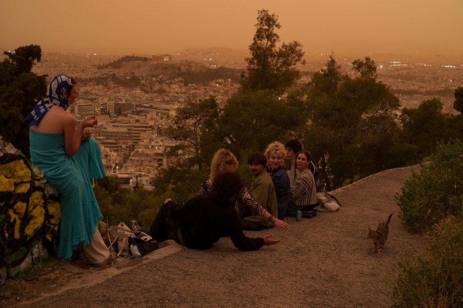 Tourists play with a cat at Lycabettus hill as the city of Athens with the ancient Acropolis hill is seen at the background, on Tuesday, April 23, 2024. The Acropolis and other Athens landmarks took on Martian hues Tuesday as stifling dust clouds blown across the Mediterranean Sea from North Africa engulfed the Greek capital. (AP Photo/Petros Giannakouris)