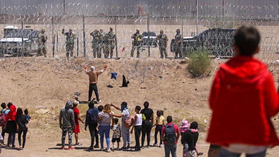 A group of migrants encounter Texas officials on the opposite side of a barbed wire fence.