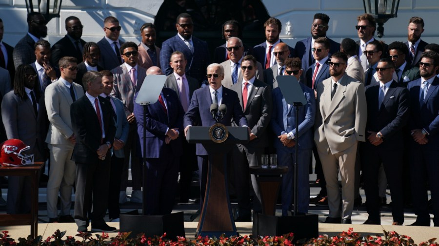 President Biden speaks while football players surround behind him.