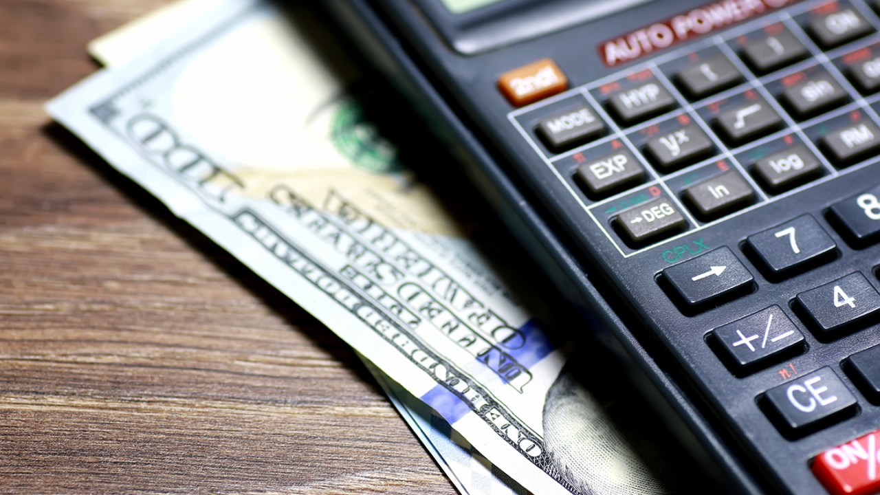 A $100 bill is visible sticking out from under a calculator in a close-up.