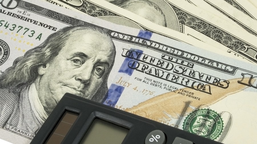 A fanned out stack of $100 bills are seen under the corner of a calculator in a close-up.