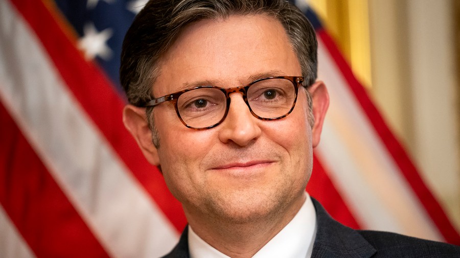 Speaker Mike Johnson smiles in a close-up in front of an American flag.