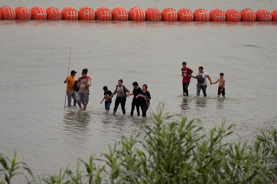 FILE - Migrants who crossed the Rio Grande from Mexico walk past large buoys being deployed as a border barrier on the river in Eagle Pass, Texas, July 12, 2023. The floating barrier is being deployed in an effort to block migrants from entering Texas from Mexico. (AP Photo/Eric Gay, File)