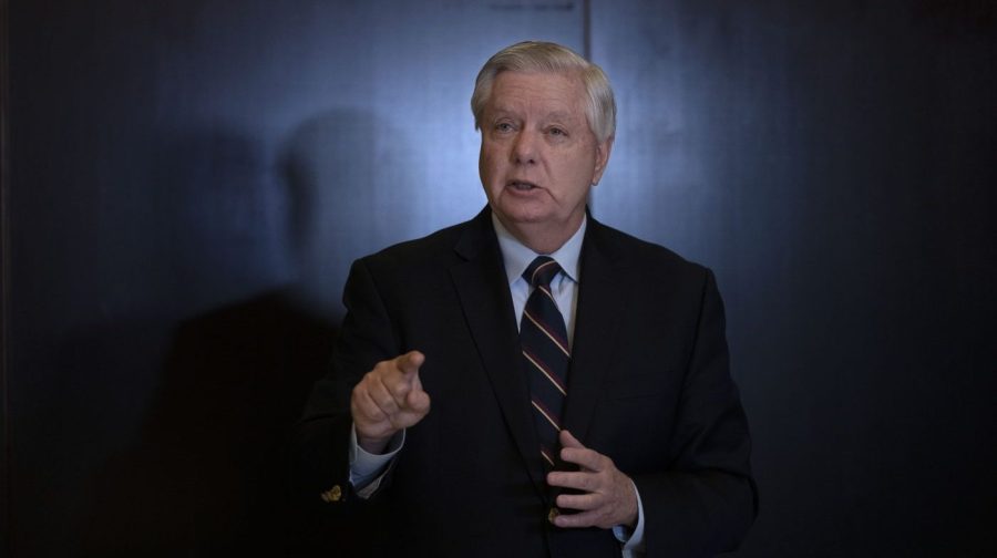 Senator Lindsey Graham gestures while speaking to the press in dim lighting.