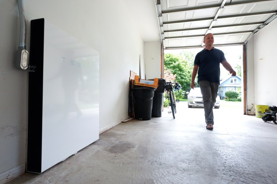 Jason Benedict walks into his garage near a Tesla Powerwall battery, left, in Berkley, Mich., Wednesday, July 24, 2024. (AP Photo/Paul Sancya)